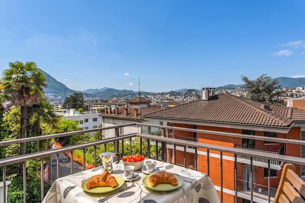 Varanda com mesa posta para café da manhã, com vista para paisagem urbana com edifícios e montanhas.