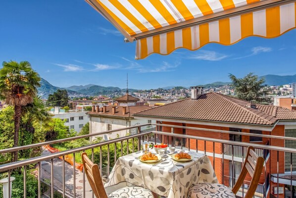 Ein Balkon mit gestreifter Markise, einem Tisch mit Croissants und Blick auf die Stadt.