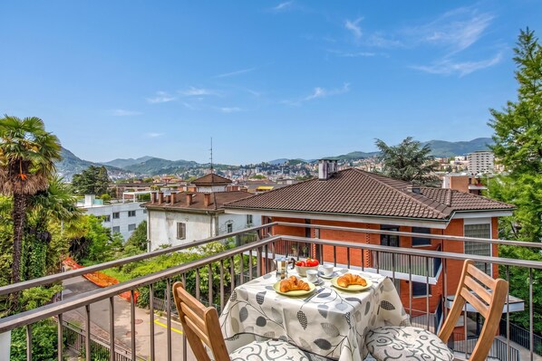 A balcony with a table set for breakfast, overlooking a cityscape with buildings and hills.