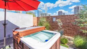 Un jacuzzi con una sombrilla roja, rodeado de un muro de ladrillo y follaje, con un horizonte urbano al fondo.
