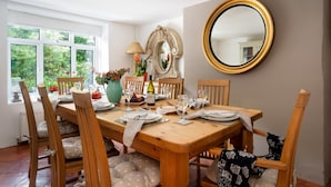 A dining room with a wooden table set for six, surrounded by wooden chairs with patterned cushions. A large mirror with a gold frame hangs on the wall, and a window provides a view of greenery outside. - Haldon View in Bishopsteignton (Teignmouth)