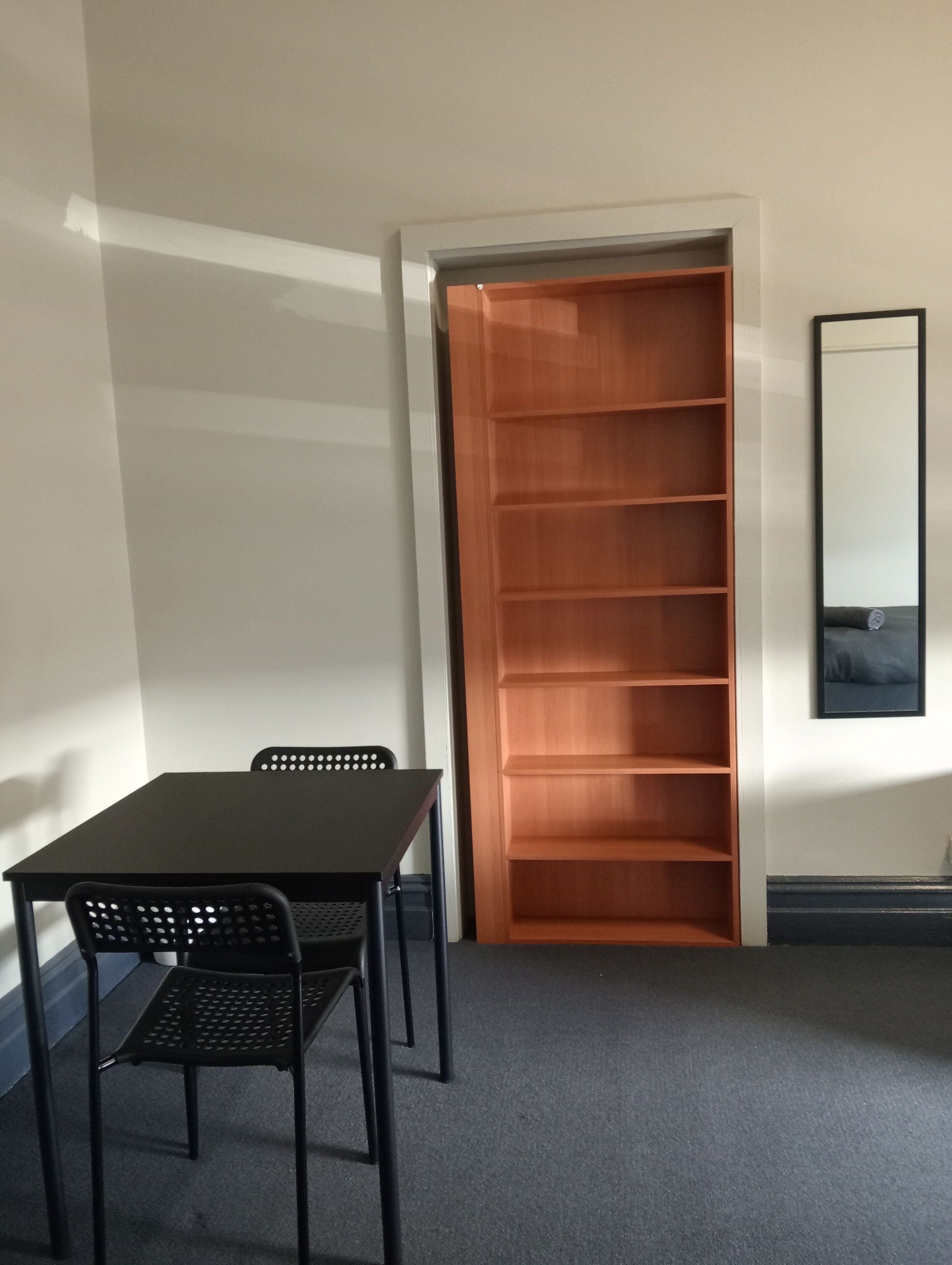 A room with a dark grey carpet, a black table with two chairs, and a wooden shelving unit against the wall.