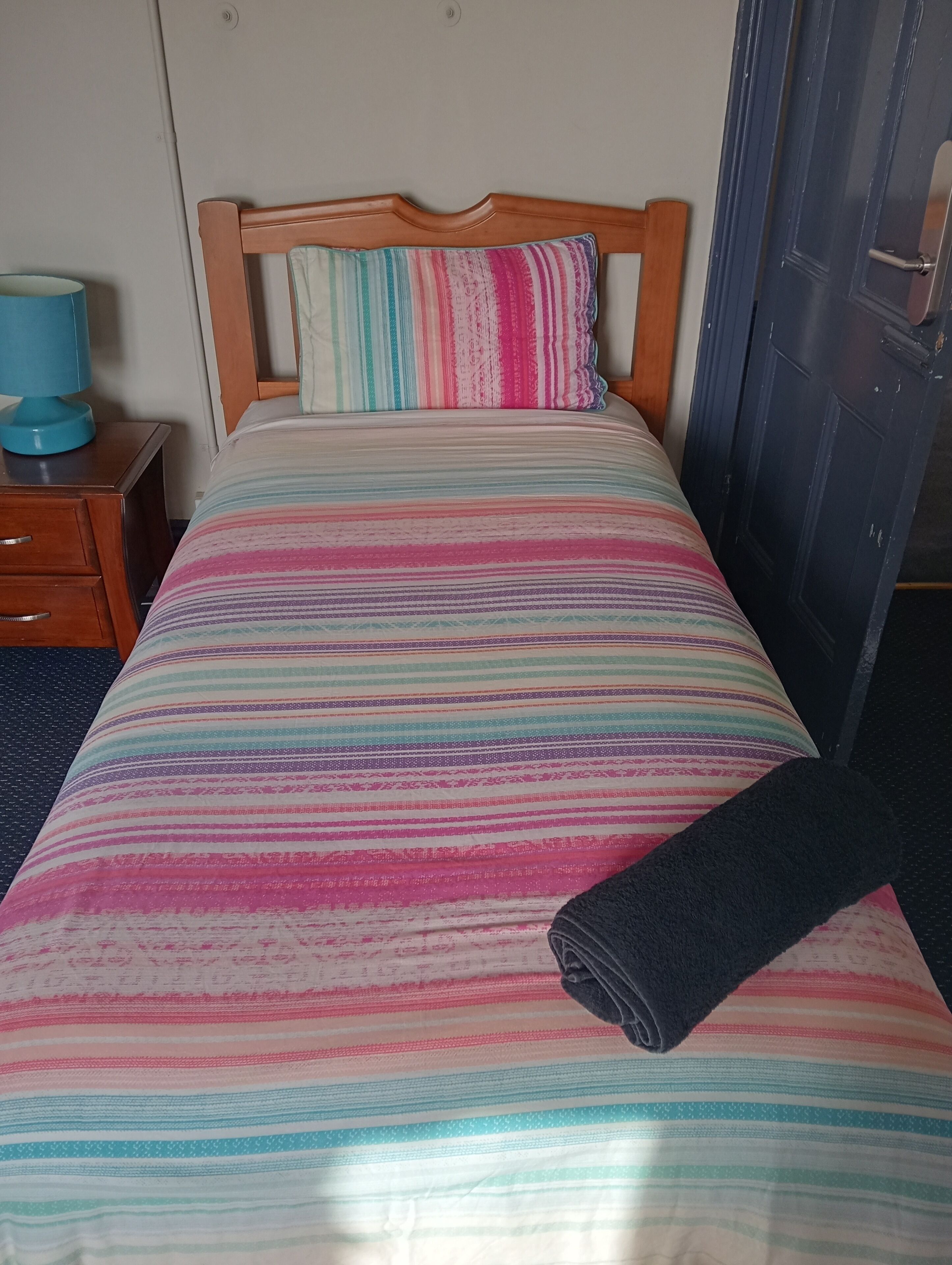 A bed with a striped bedspread, a wooden headboard, a blue lamp, and a dark-colored blanket on the floor.