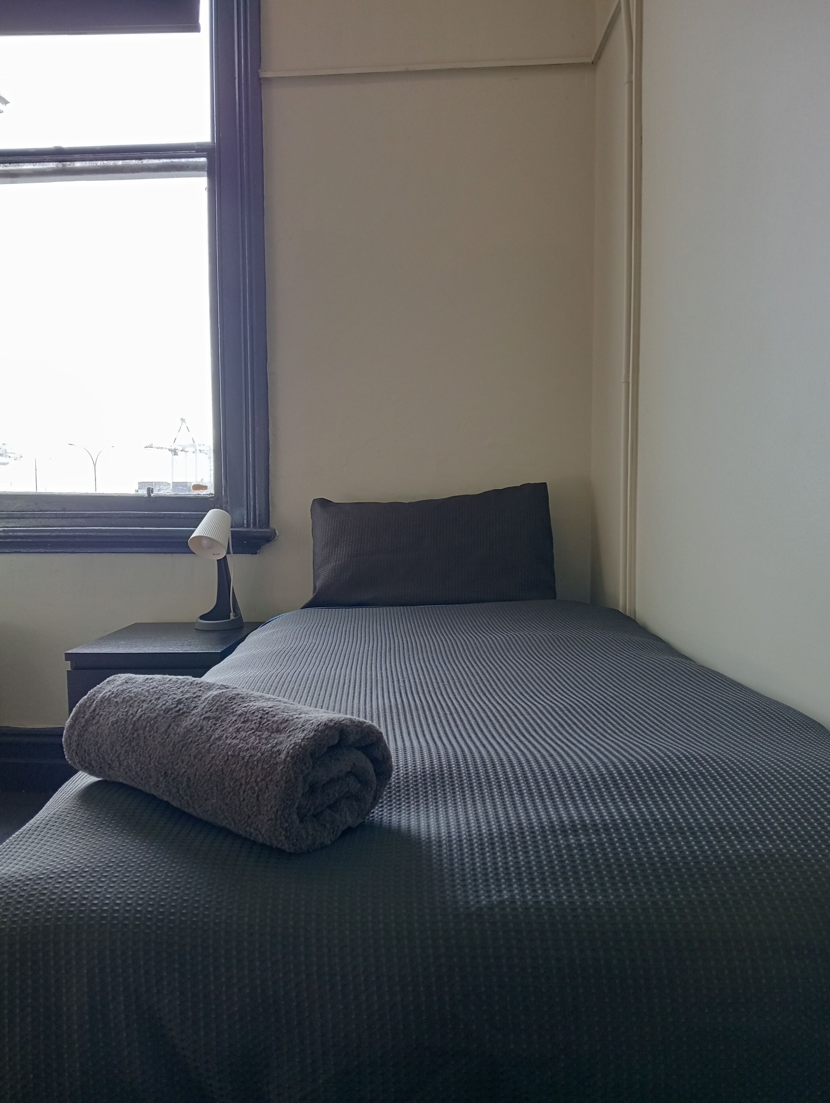 A neatly made bed with a dark gray sheet and a rolled towel on top. There is a single dark gray pillow and a small bedside table with a lamp. A window with a view of construction cranes is visible in the background.