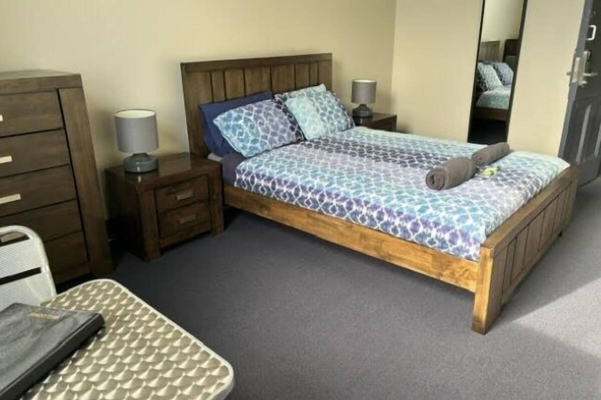 Un lit simple avec une couvre-lit à motifs, une tête de lit en bois, une table de chevet avec une lampe, un miroir et un bureau avec une chaise.