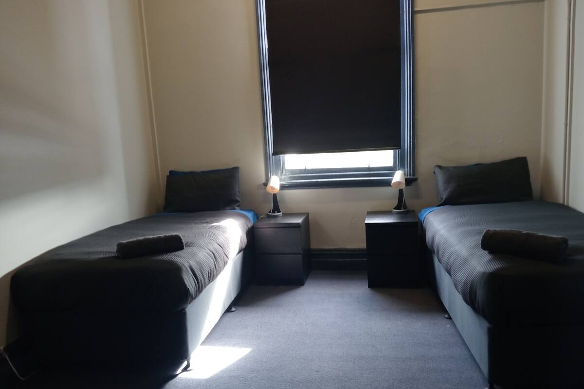 Une chambre avec deux lits simples, chacun étant équipé d’un oreiller et d’une table de chevet avec une lampe. Il y a une fenêtre au-dessus des lits.
