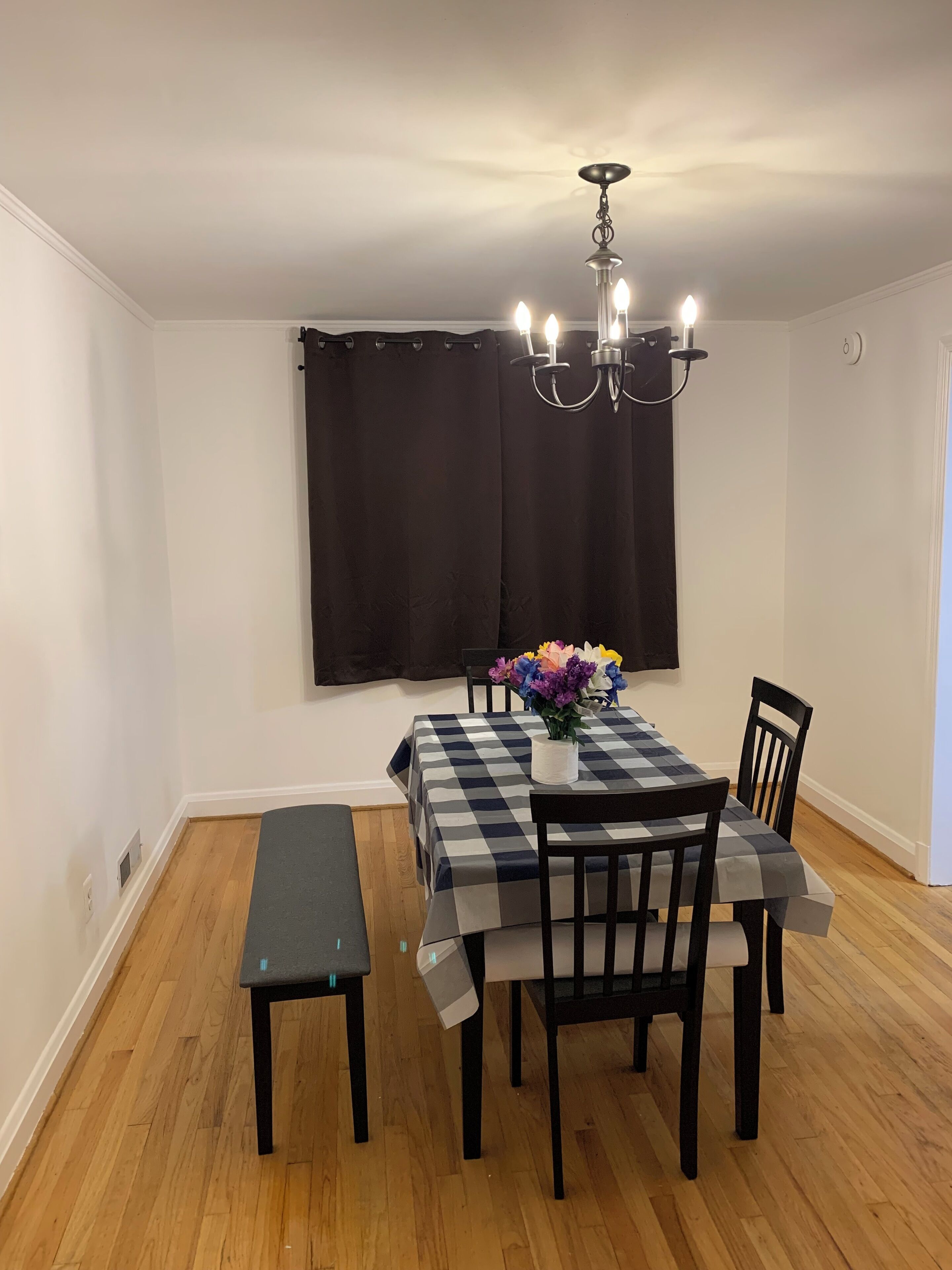 A dining area with a checkered tablecloth, black chairs, a bench, a chandelier, and a window with curtains.