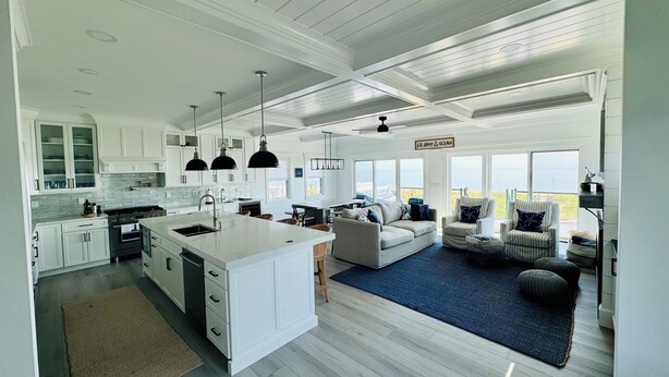 Une cuisine spacieuse avec des meubles blancs, un grand îlot central et des luminaires suspendus. On y trouve un coin salon confortable avec un canapé et des fauteuils, et une vue sur l’océan grâce à de grandes fenêtres.