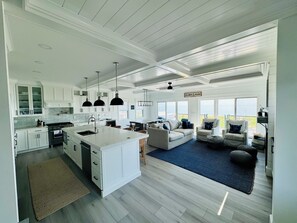 Una cocina amplia con armarios blancos, una isla grande e iluminación colgante. Hay una zona de estar acogedora con un sofá y sillones, y vistas al océano a través de grandes ventanales.