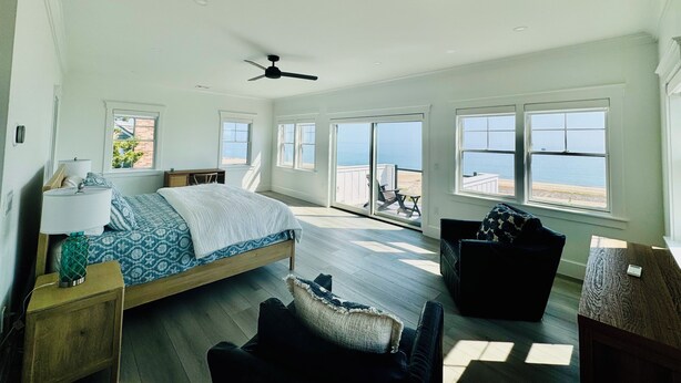 Un dormitorio amplio con una cama grande, ventilador de techo y varias ventanas con vista a la playa.