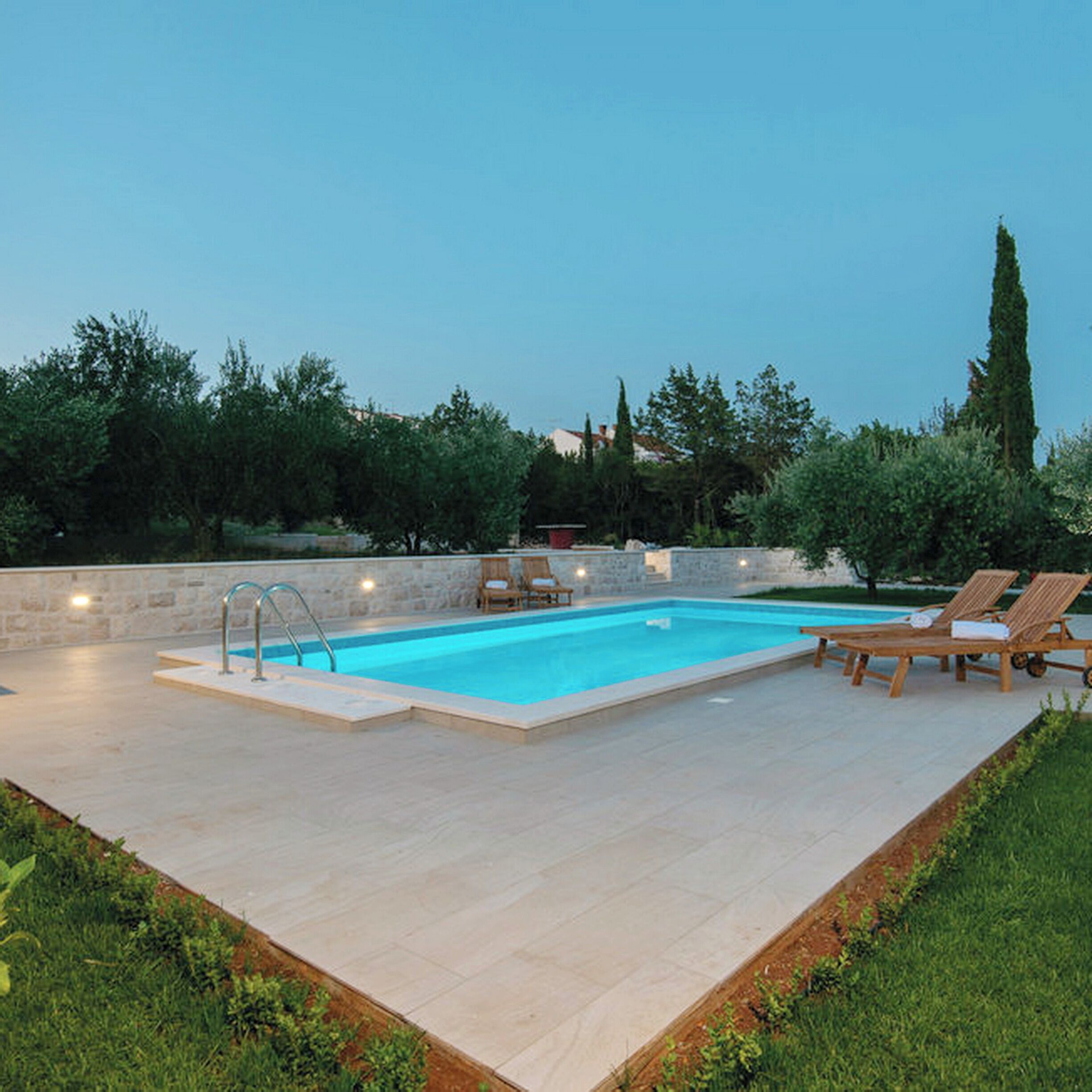A rectangular swimming pool with a stone deck, surrounded by lounge chairs and a low stone wall.