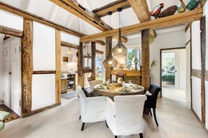 A dining area with a round table, white upholstered chairs, hanging pendant lights, and exposed wooden beams.
