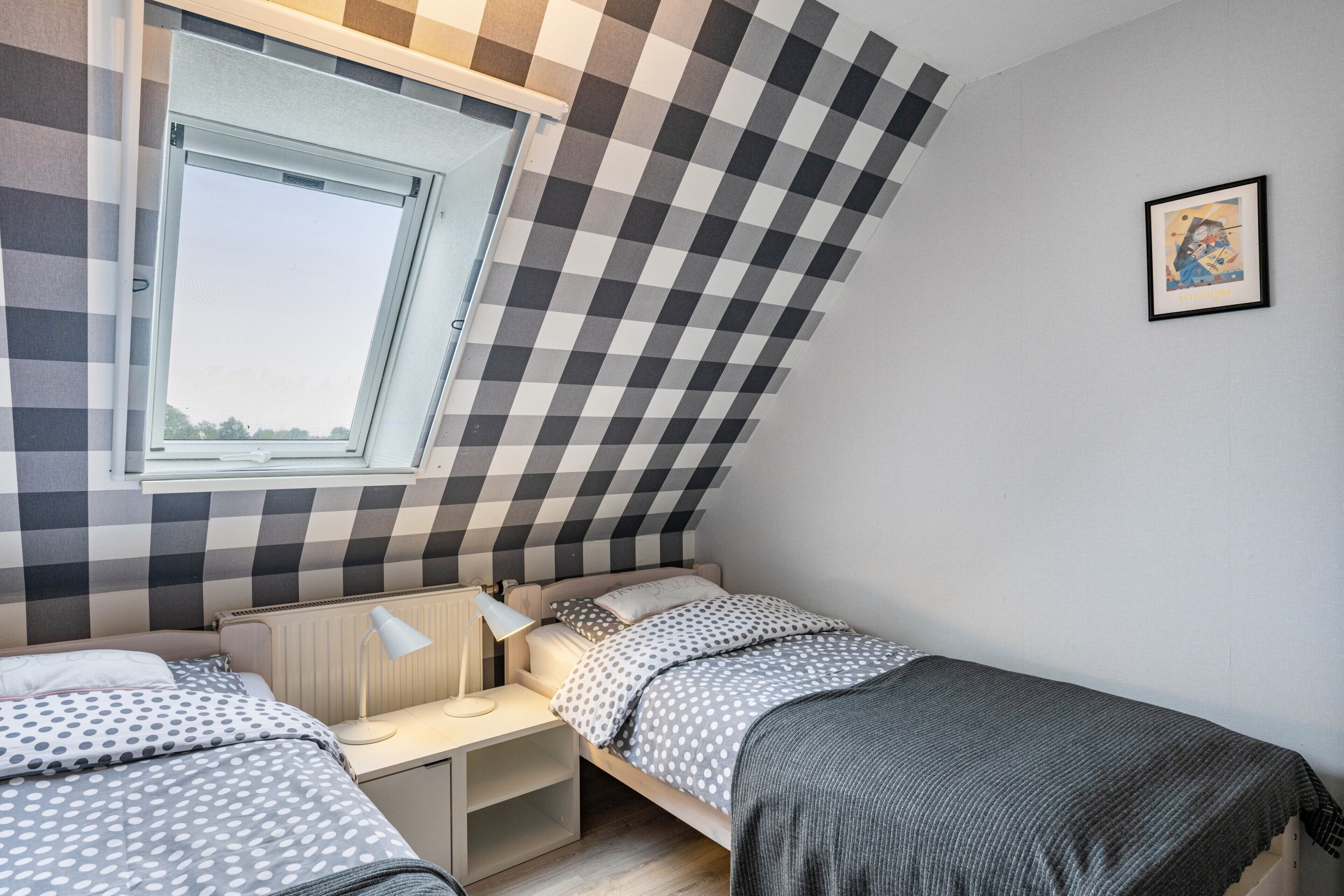 A bedroom with a checkered wall, a window, a bed, a desk, and a lamp.