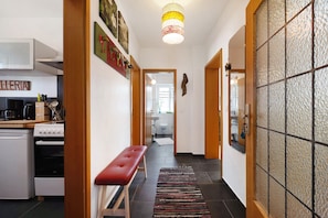 Un couloir étroit avec un banc, un coin cuisine équipé d’appareils électroménagers et un mur recouvert de carreaux.