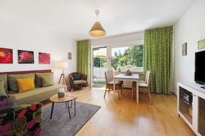 Ein Wohnzimmer mit einer Couch, einem Esstisch mit Stühlen, einem Fernseher und einem Fenster mit Vorhängen.
