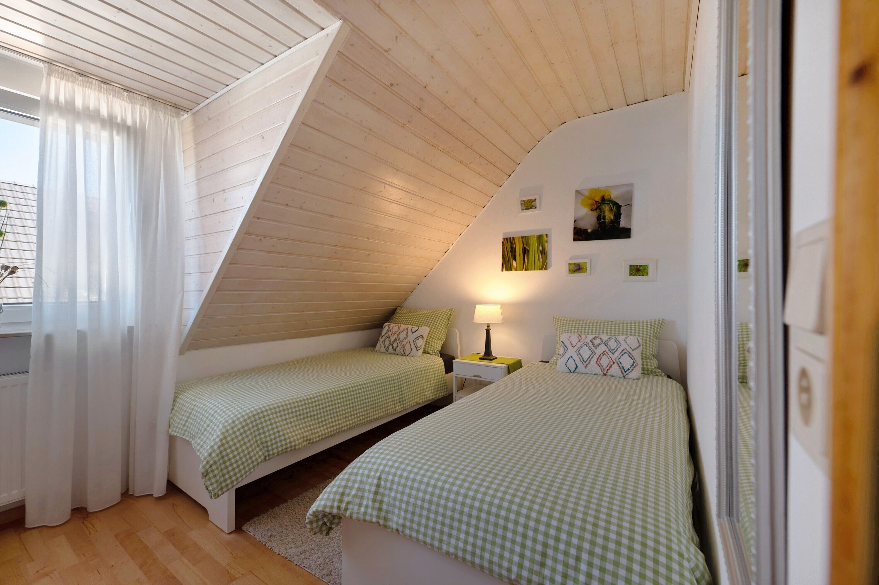 Een slaapkamer met twee eenpersoonsbedden, een schuine plafond, een raam met gordijnen, een nachtkastje met een lamp en aan de muur hangende foto's.