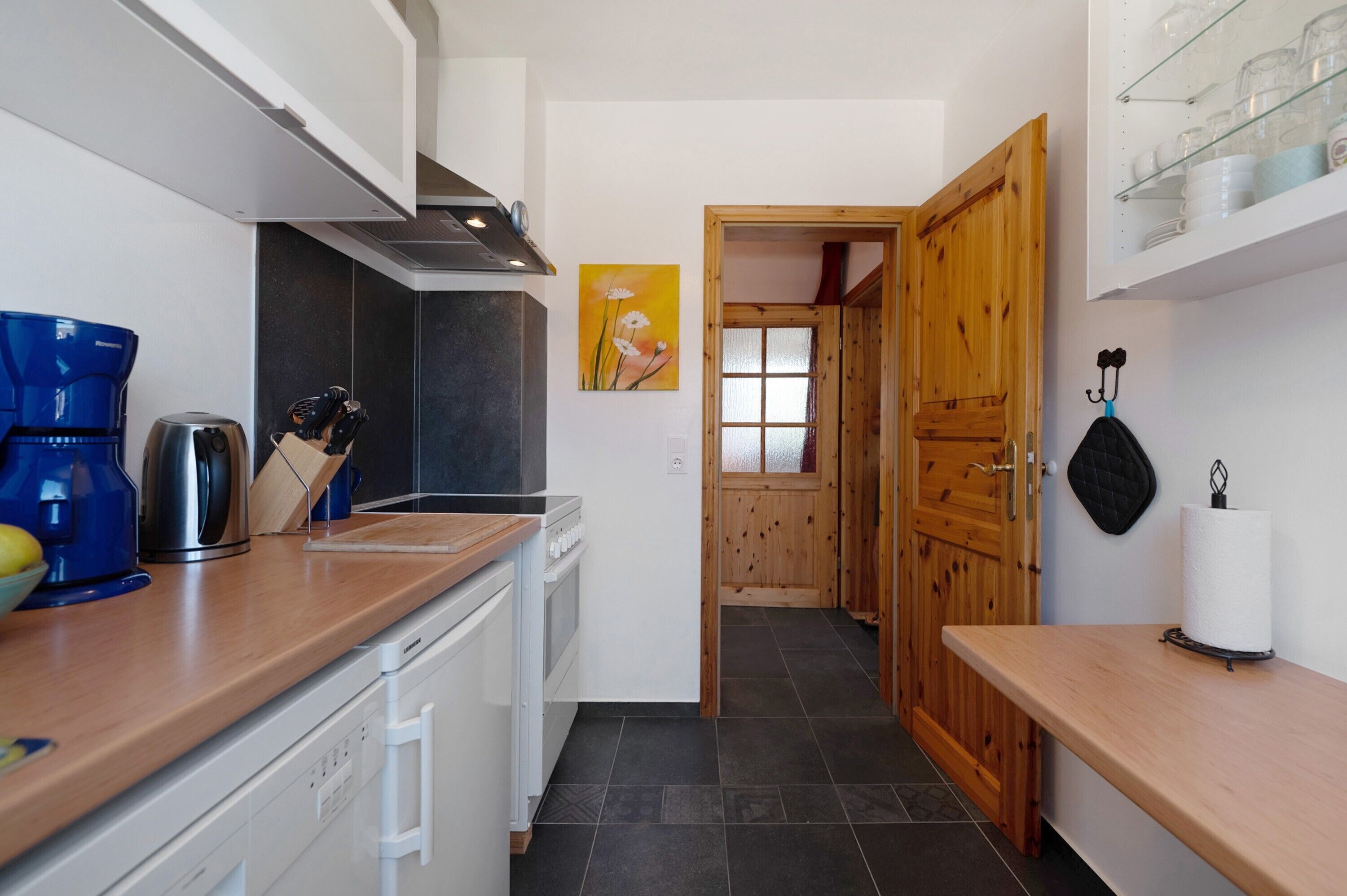 Une cuisine équipée d’une machine à café, d’une bouilloire et d’ustensiles de cuisine posés sur le plan de travail, une porte en bois donnant sur une autre pièce, et un étagère fixée au mur pour ranger la vaisselle.