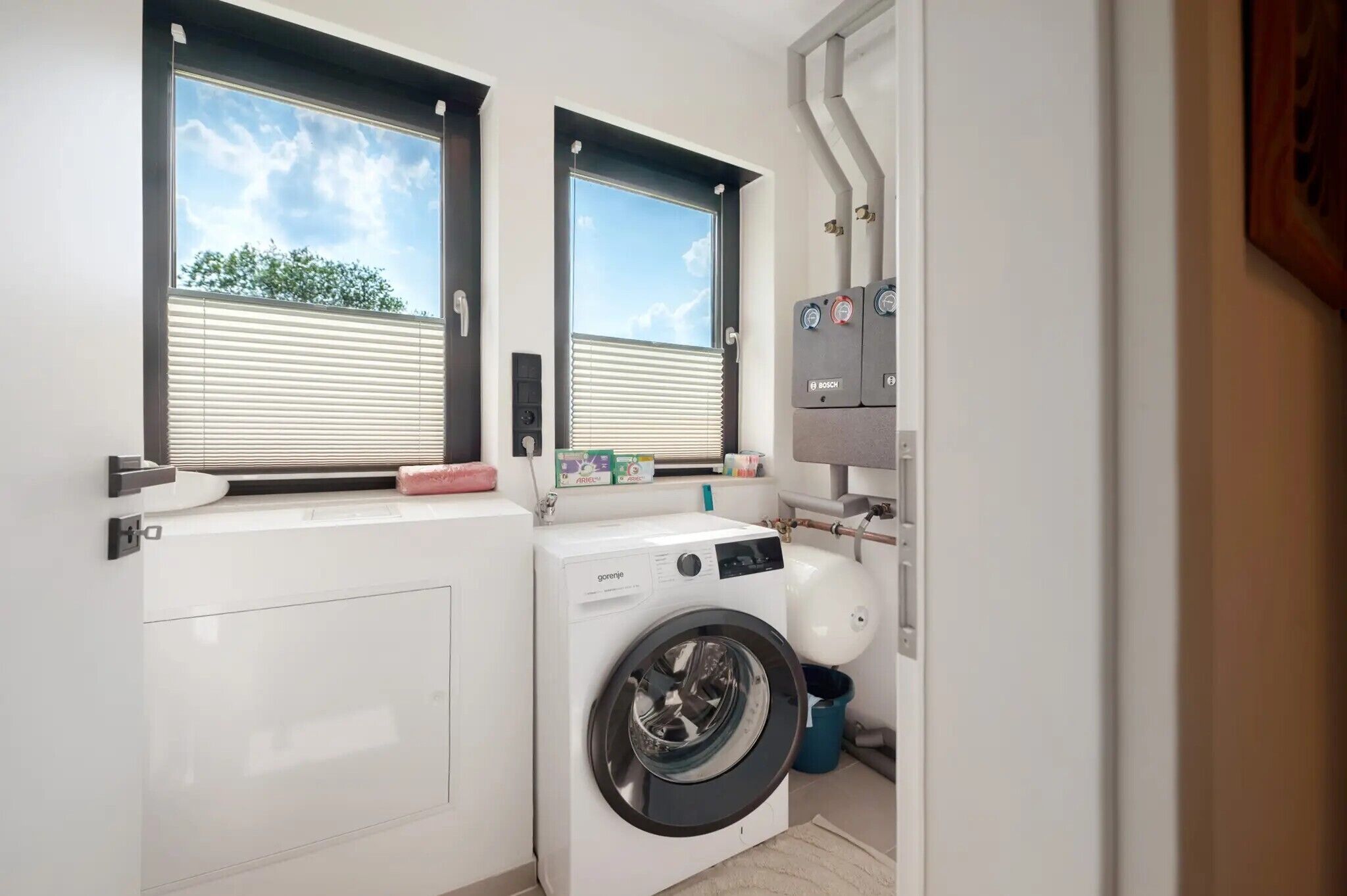 Une petite buanderie équipée d’un lave-linge, d’un sèche-linge et d’une fenêtre avec des stores.