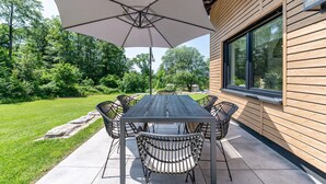 An outdoor patio with a table and chairs, a large umbrella, and a view of a wooden building with a window.