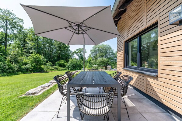 Eine Terrasse im Freien mit Tisch und Stühlen, einem großen Sonnenschirm und Blick auf ein Holzbauten mit einem Fenster.