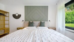 A bedroom with a bed, two bedside tables, a wall-mounted light fixture, a wooden shelf, and a window with a view of greenery.