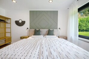 Ein Schlafzimmer mit einem Bett, zwei Nachttischen, einer Wandleuchte, einem Holzregal und einem Fenster mit Blick ins Grüne.