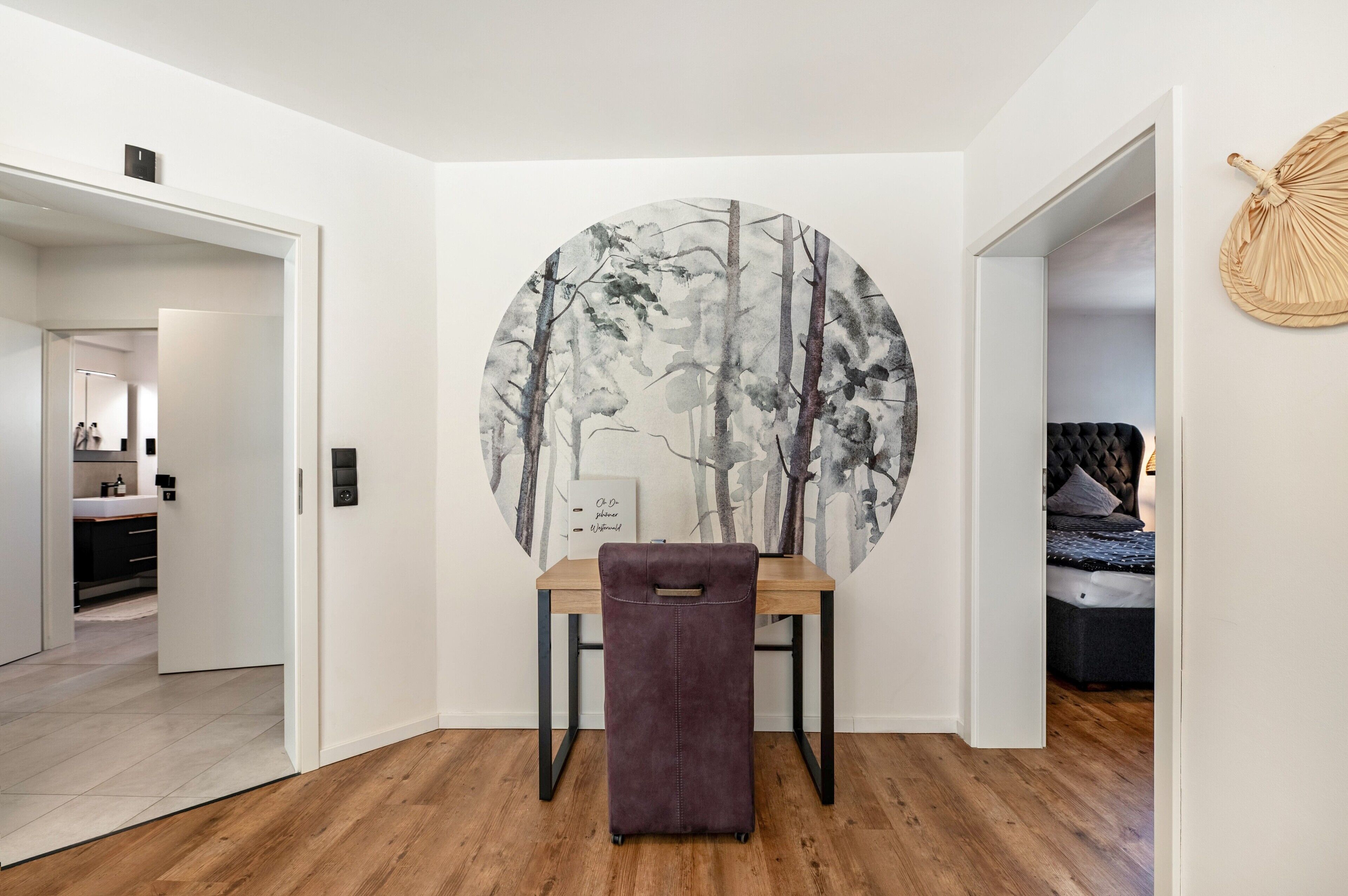 Una habitación con un escritorio de madera, una silla, un cuadro circular grande con un diseño de árboles, una cama y un tapiz de pared tejido.