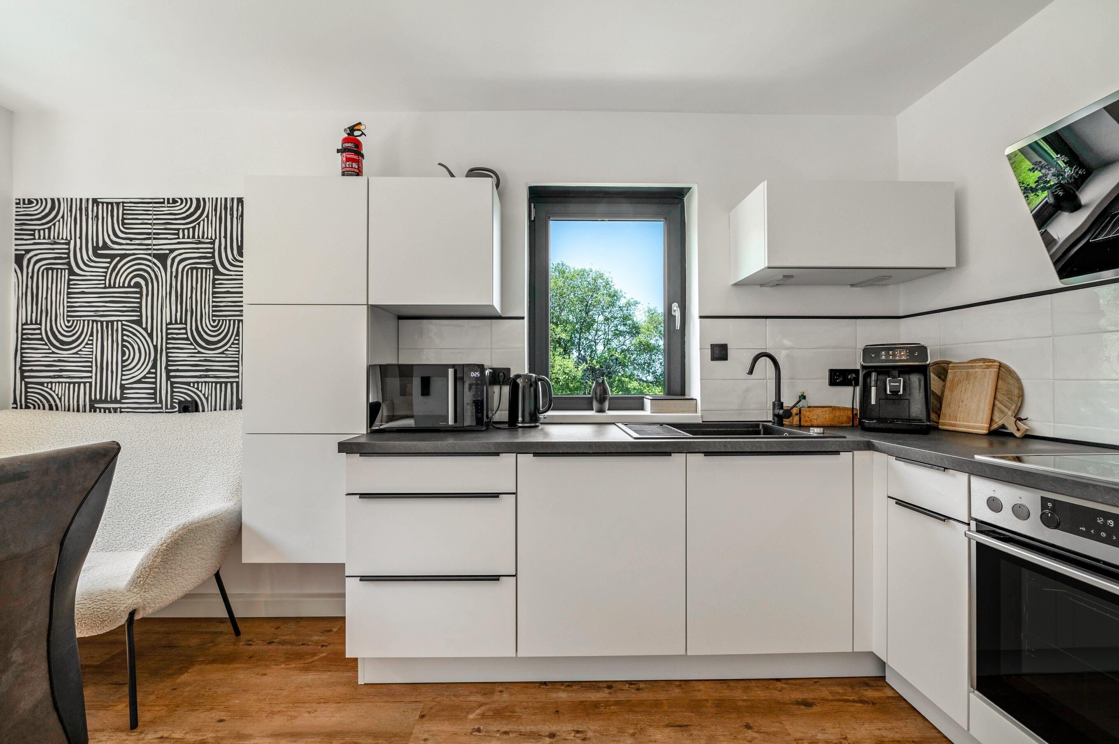 A modern kitchen with white cabinets, a black countertop, a window with a view of trees, and a small stove.