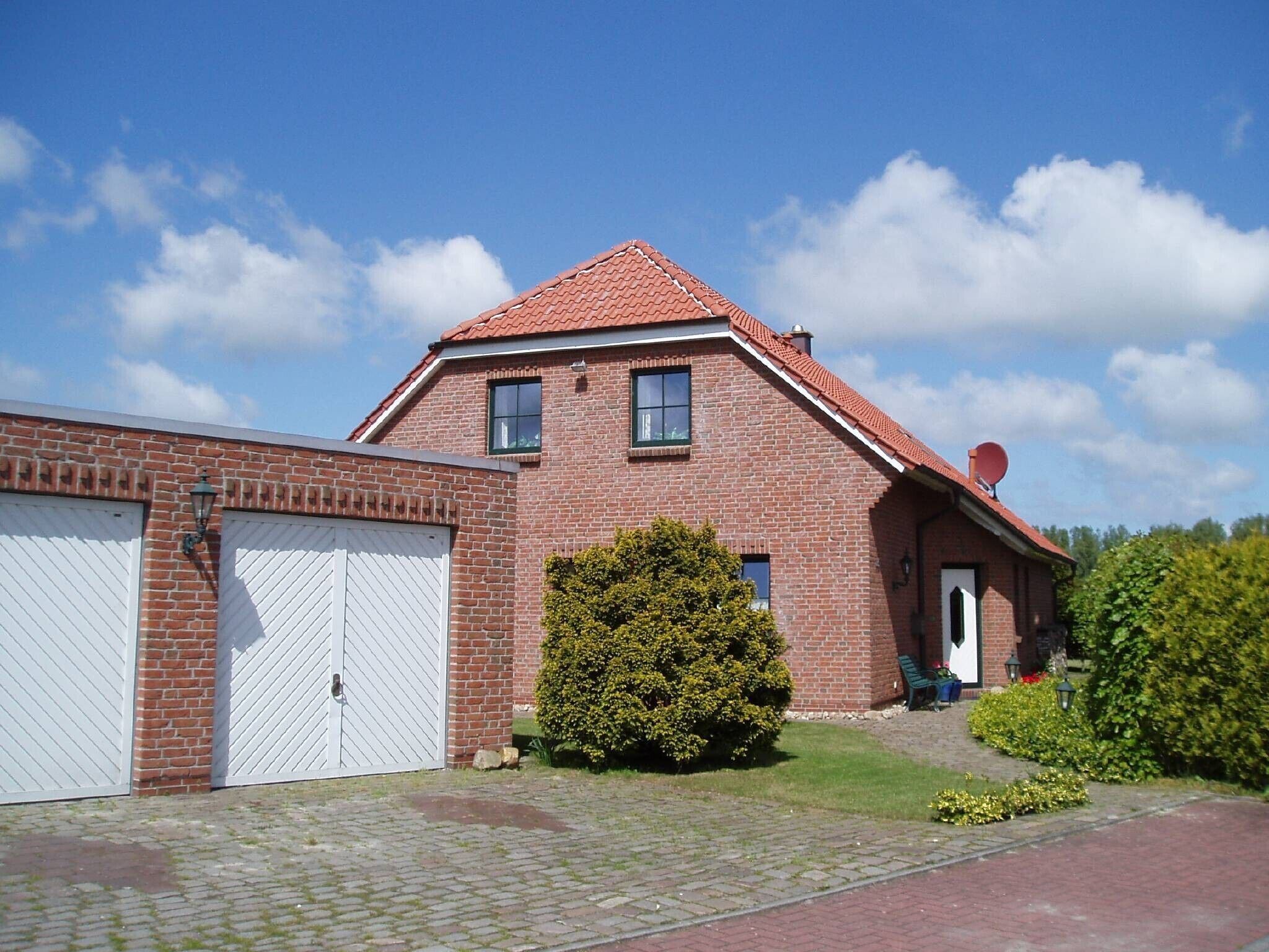 A two-story brick house with a red-tiled roof, a white garage door, and a small front yard with a bush.