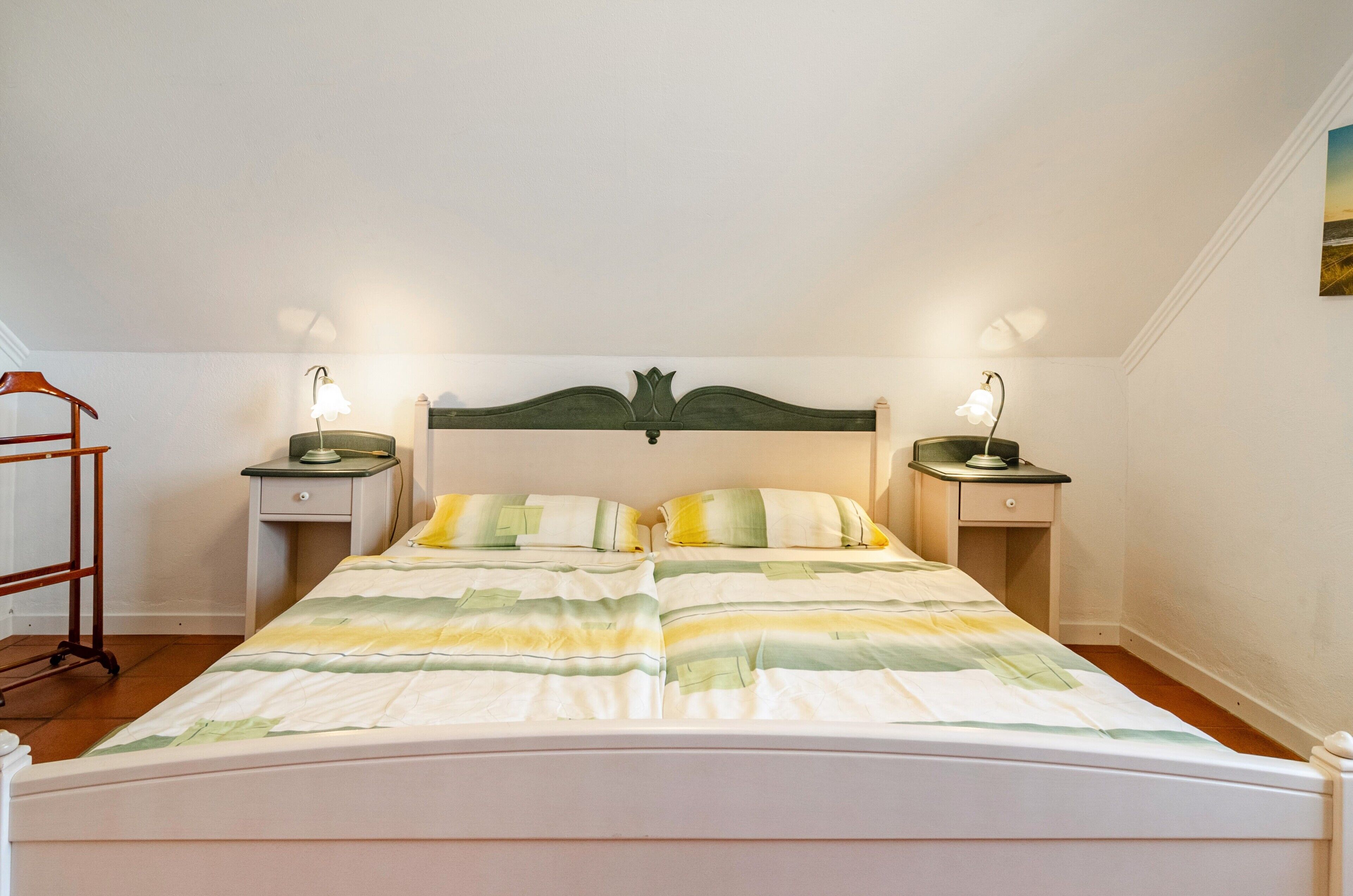 A bedroom with a bed, two bedside tables, a nightstand, a lamp, and a wall-mounted shelf.