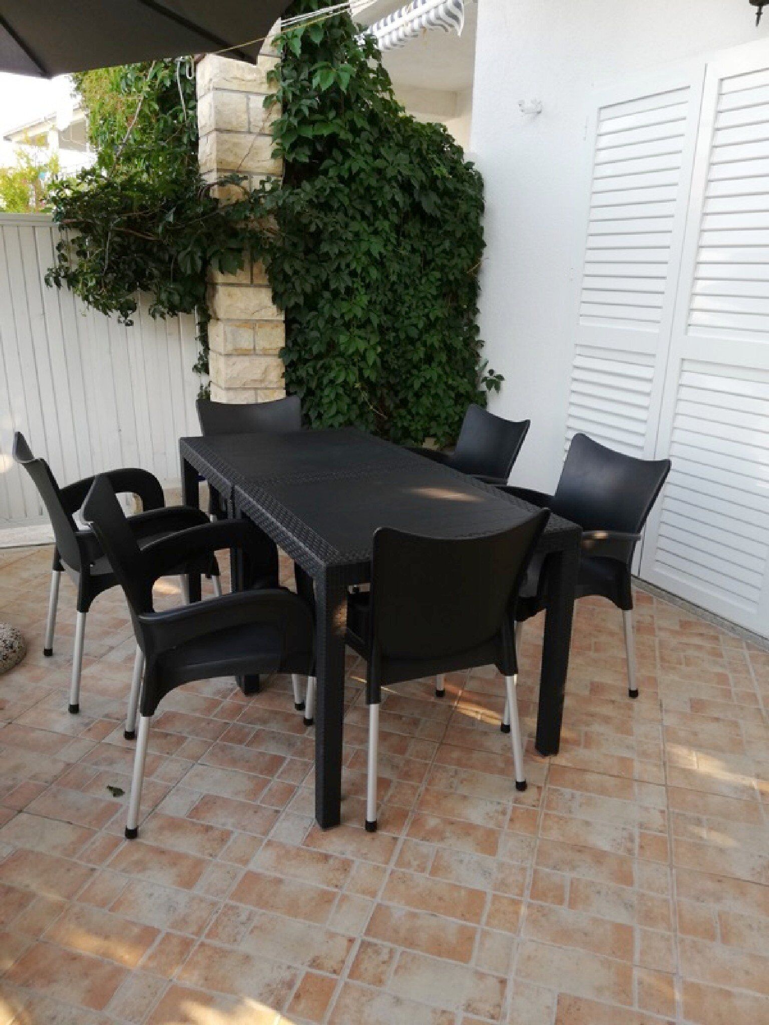 En udendørs terrasse med et sort bord og stole, omgivet af et hvidt hegn og en stenvæg med grøn vegetation.