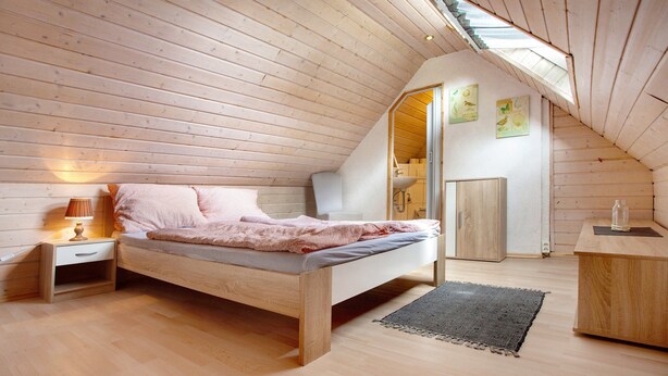 Un dormitorio con techo inclinado de madera, una cama con manta rosa, una mesita de noche con lámpara y un escritorio con una taza.