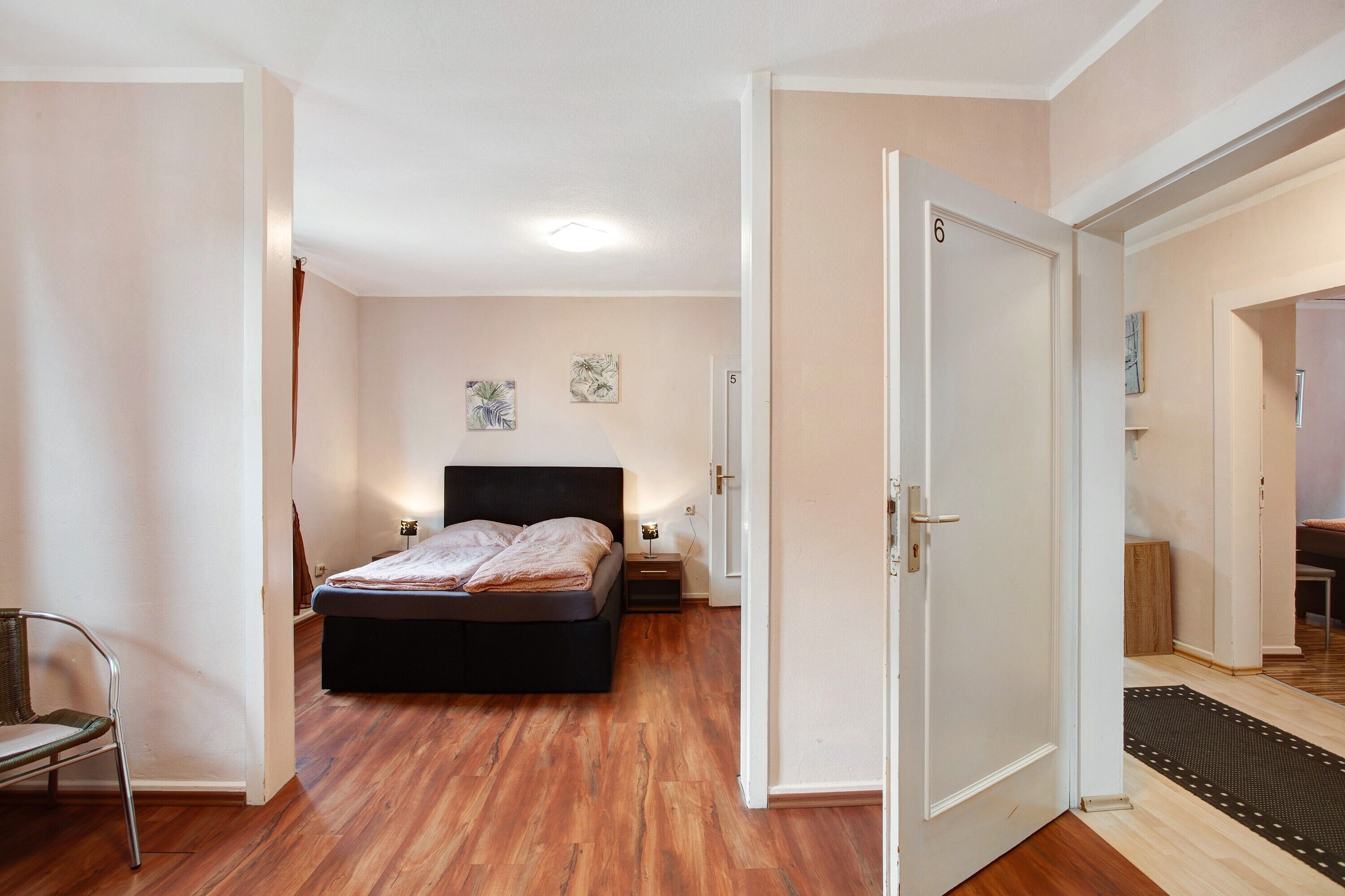 Ein Schlafzimmer mit einem Bett, Nachttischen, einem Sessel und einer Tür zu einem anderen Raum.