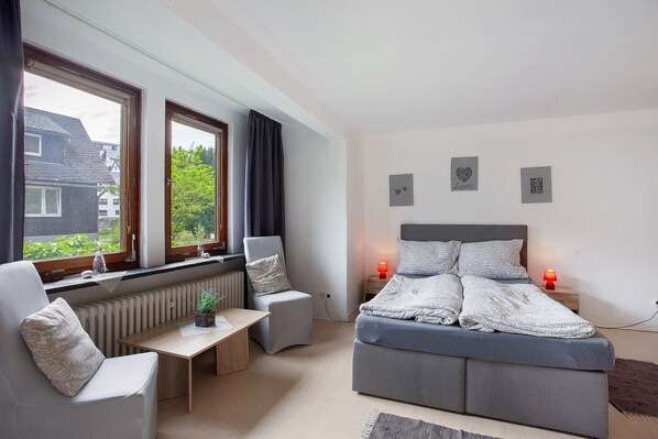 A bedroom with a bed, two bedside tables with lamps, a chair, a radiator, and a window with curtains.