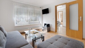 A modern living room with a sofa, a glass coffee table, a flat-screen TV mounted on the wall, and a sliding door leading to another room. - Holiday Home 'Ferienhaus' with Private Terrace, Shared Garden and Wi-Fi (Großkoschen)