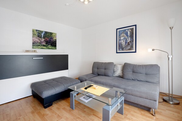 A modern living room with a grey sofa, a glass coffee table, a bench, a TV, and two framed artworks on the wall.