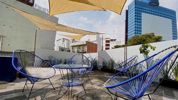 Een terras buiten met blauwe draadstoelen en een tafel, een groot zonnescherm en een uitzicht op moderne gebouwen op de achtergrond.
