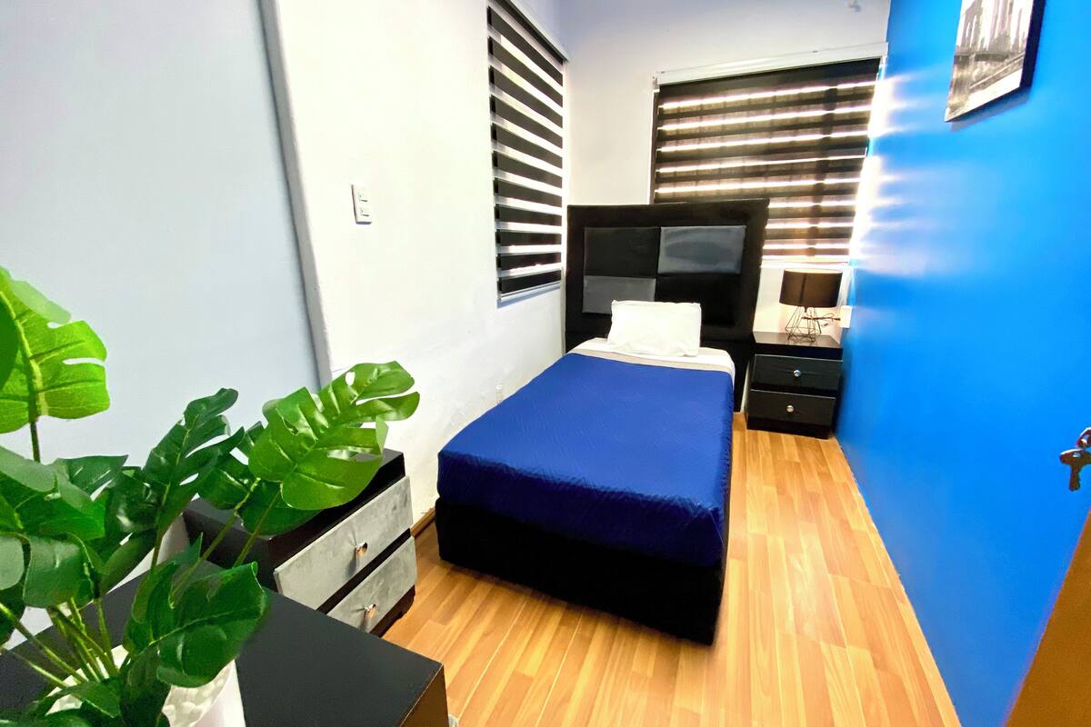 Een slaapkamer met een bed, nachtkastje, dressoir, een potplant en een raam met jaloezieën.