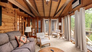 Ein Interieur einer Holzblockhütte mit Holzbalkendecke, Deckenventilator, einer Couch, einem Couchtisch und Blick ins Freie durch große Fenster.
