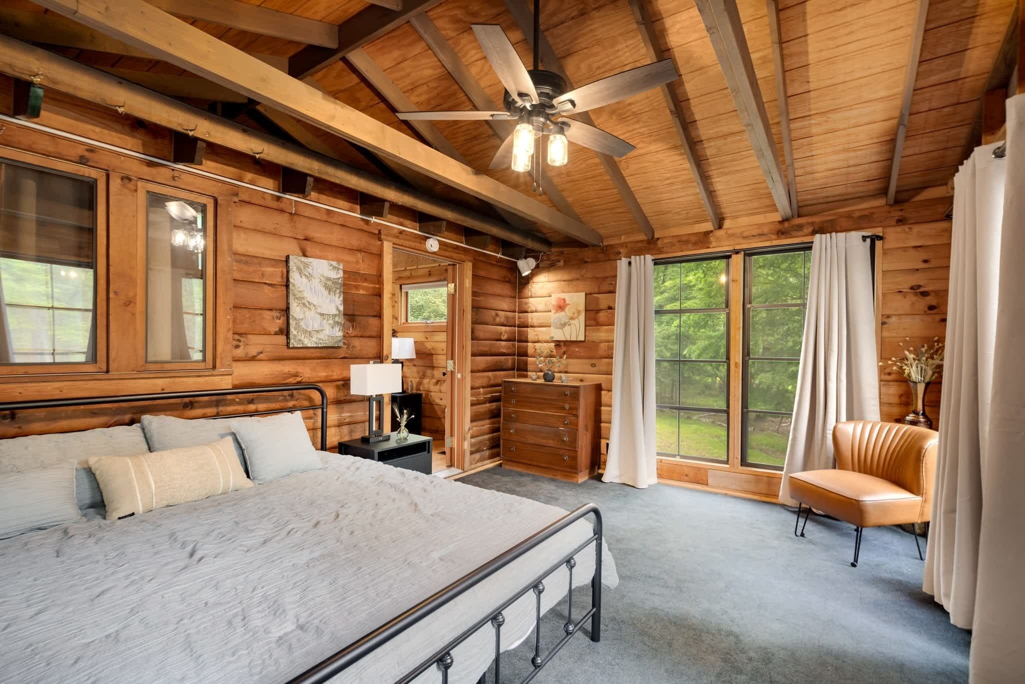 A log cabin bedroom with a bed, dresser, chair, and large windows.