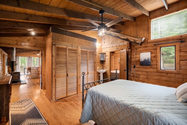 A wooden cabin interior with a bed, a ceiling fan, a sink, and a view of the outdoors. - Hot Tub, Firepit, Grill, Wifi near Golf & Hiking (Berkeley Springs)