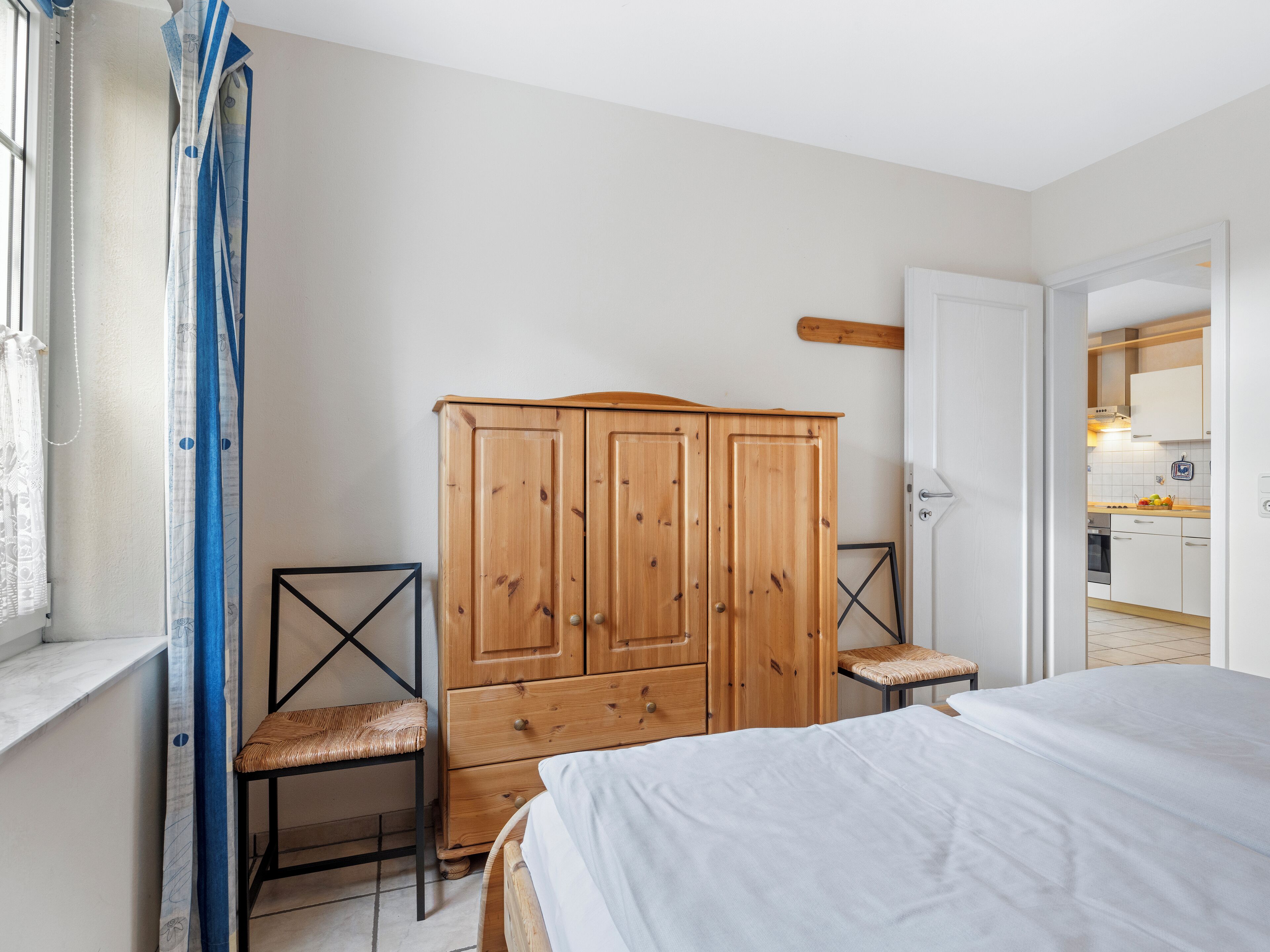 A bedroom with a wooden wardrobe, a bed, a chair, and a view of a kitchen through an open door.
