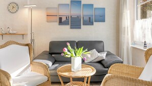 Ein modernes Wohnzimmer mit einem grauen Sofa, Korbstühlen, einem runden Tisch mit Blumenvase und einer Wand mit einer Uhr und Sonnenuntergangsbildern.