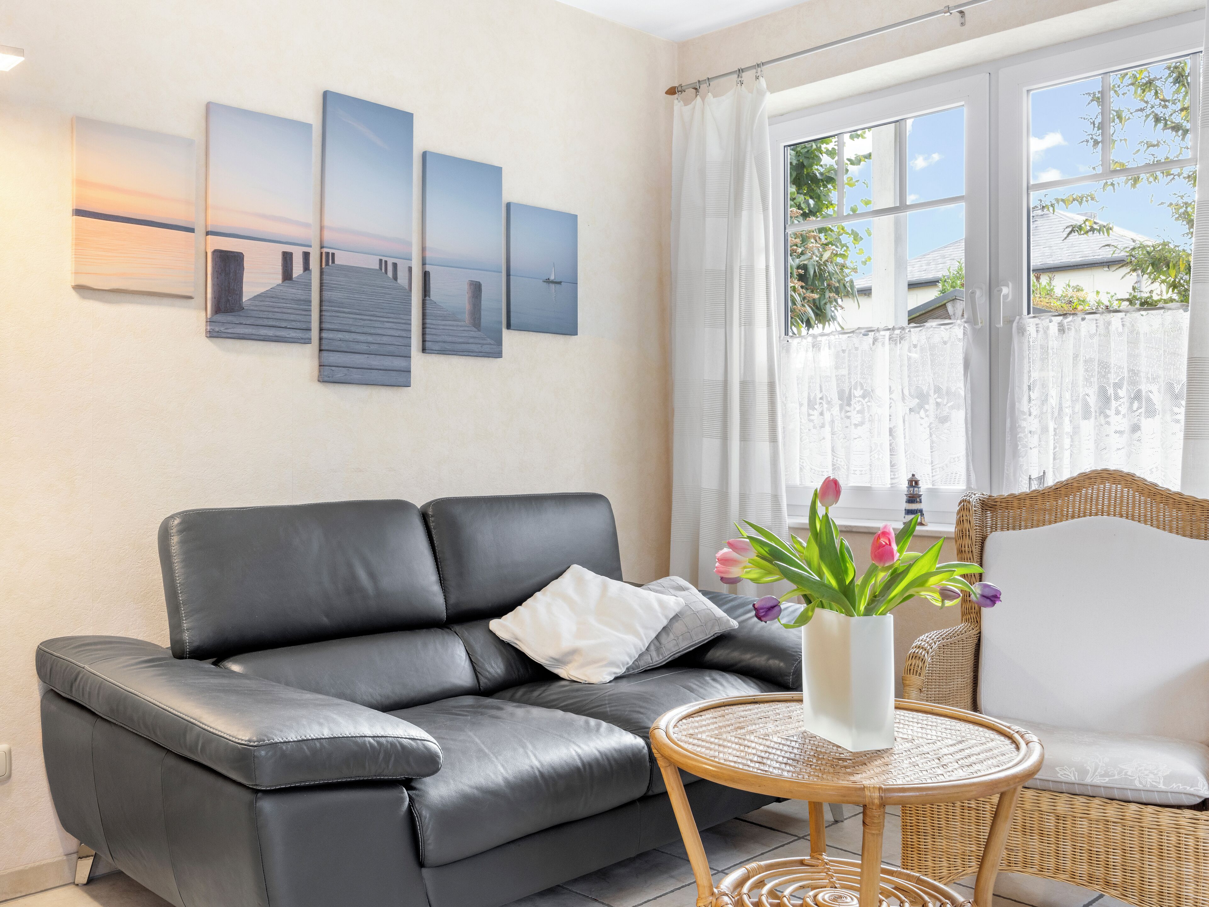 A modern living room with a black leather sofa, a round wicker table with a vase of flowers, and a view of the sea through large windows.