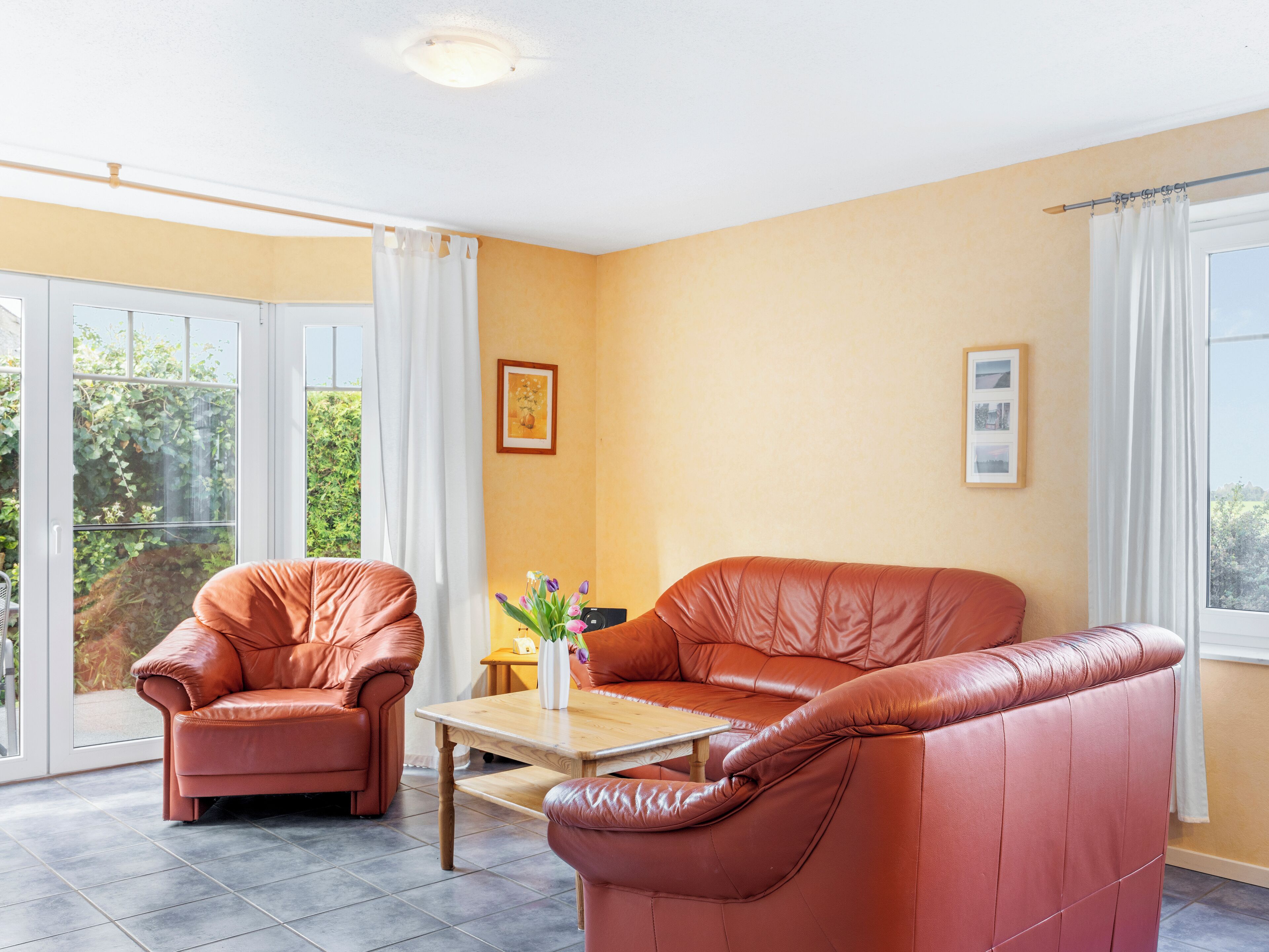 Un salon avec un canapé en cuir marron, un fauteuil en cuir marron et une table basse en bois. Il y a une fenêtre avec des rideaux blancs, un tableau encadré au mur et un vase avec des fleurs sur la table.