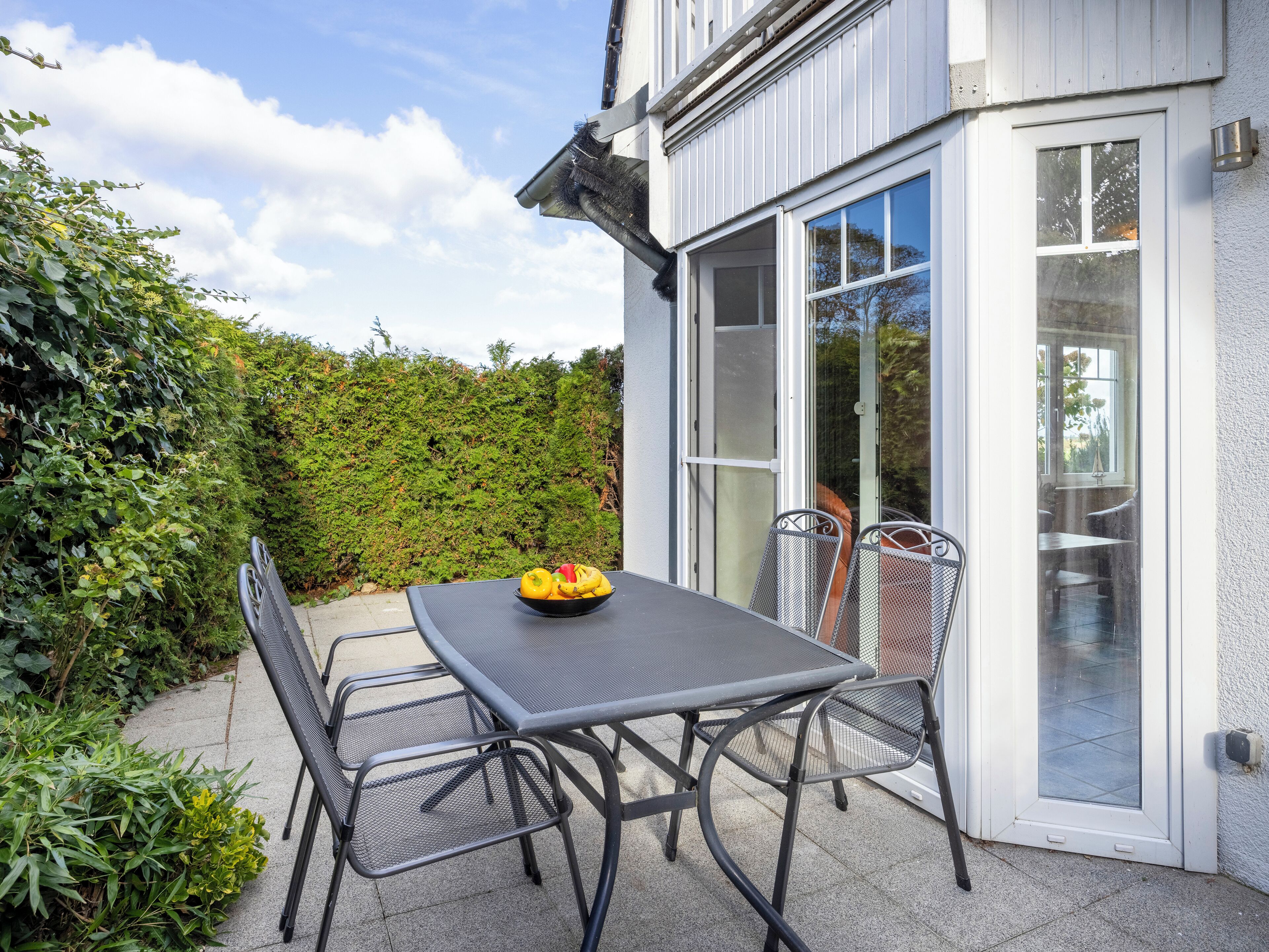 An outdoor patio with a table and chairs, a bowl of fruit, and a view of a hedge and building.