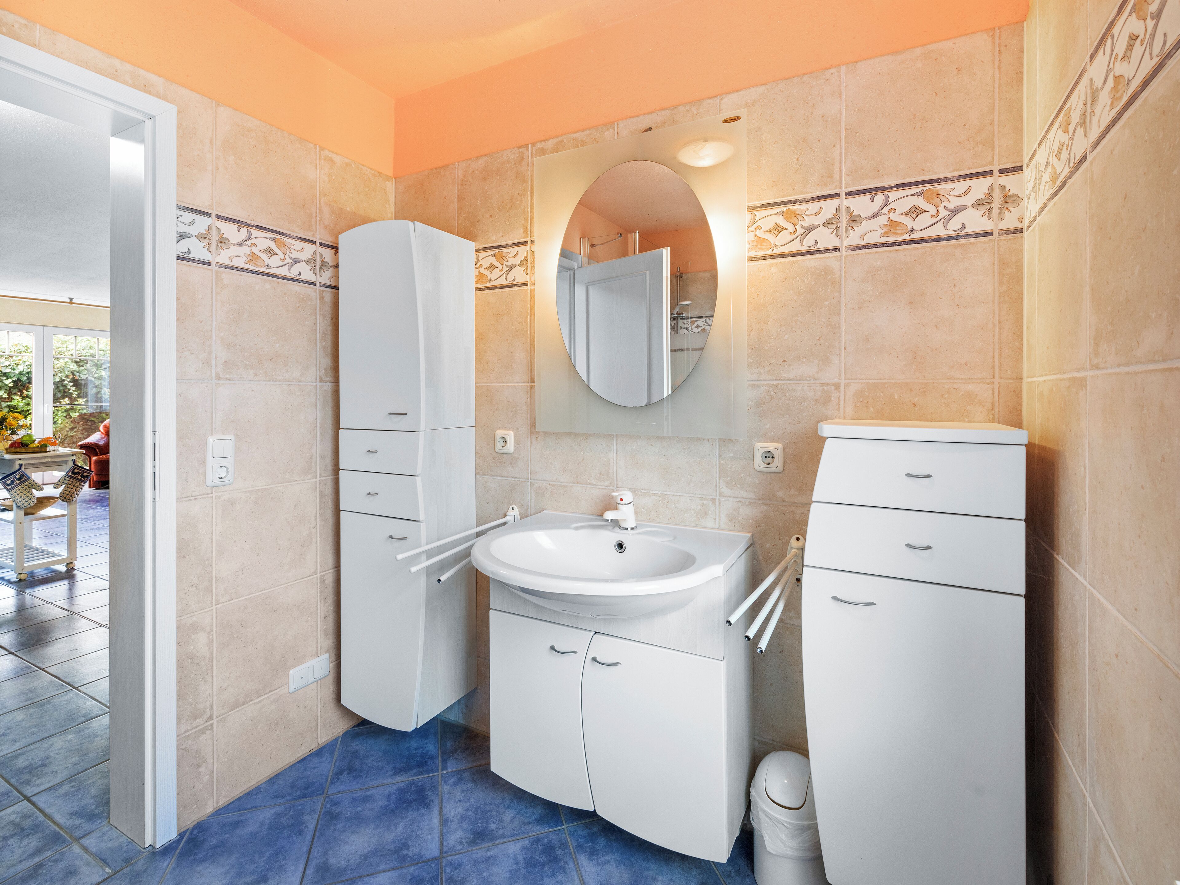 Une salle de bain avec un lavabo blanc, un miroir et des armoires, un sol carrelé de bleu et des murs de couleur beige.