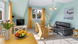 Un salon avec un canapé noir, une table à manger ornée de fruits, un téléviseur et une fenêtre avec des rideaux.