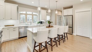 Une cuisine moderne avec des meubles blancs, un îlot central doté de tabourets, des appareils en acier inoxydable et un éclairage suspendu.