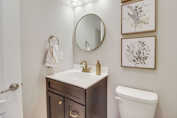 Une salle de bain avec un lavabo blanc, un meuble brun foncé, un robinet doré et des toilettes blanches. Des reproductions botaniques encadrées ornent le mur.